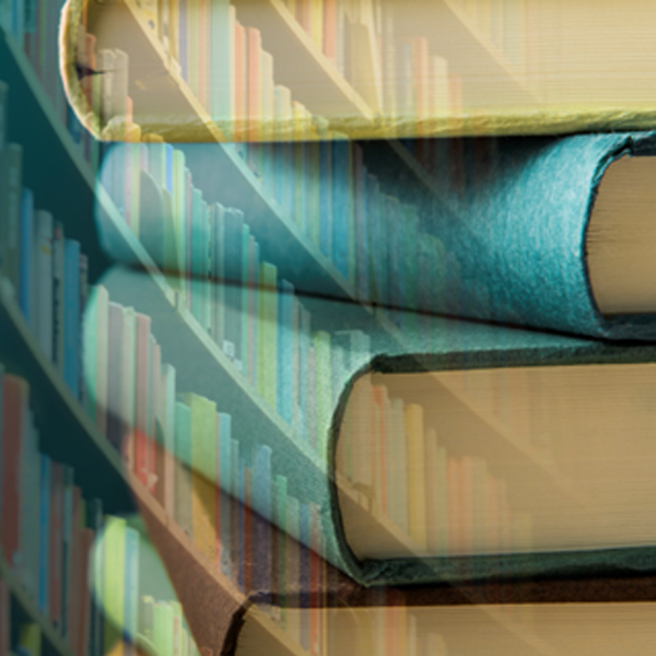 A stack of books sits in a library, where a curved wall of books can be seen in the background.